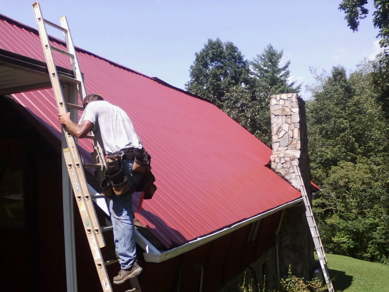 Roofer on a ladder beside a red metal roof and stone chimney — Soffit & Fascia Repair by Roofing Brothers in Richwood
